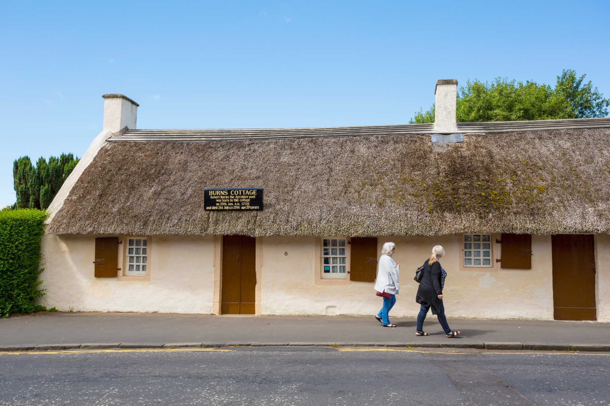 Two people walk by a thatched cottage bearing a sign that this is the birthplace of Robert Burns.